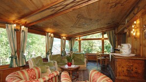 Una habitación con vigas de madera en el techo, sillones tapizados en verde, una mesa de centro de madera y vistas a árboles a través de amplias ventanas.