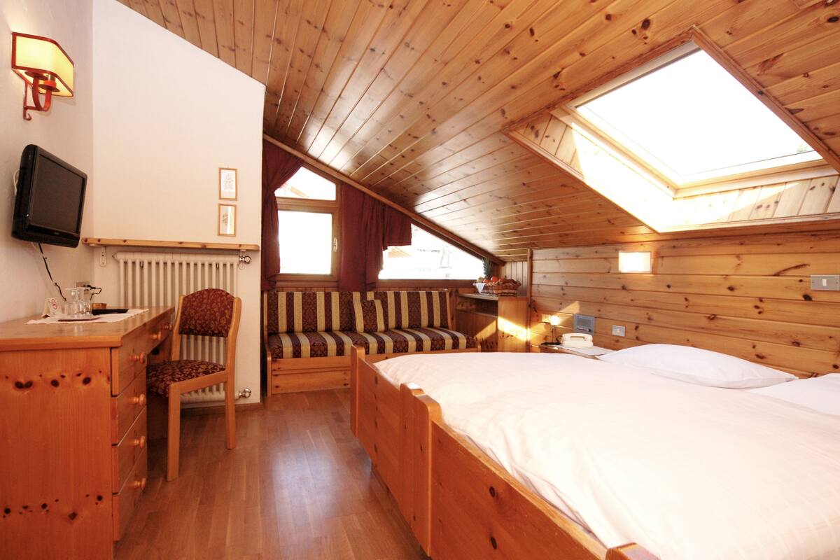 A cozy room with a wooden ceiling, a bed, a bench, a desk with a chair, and a television.