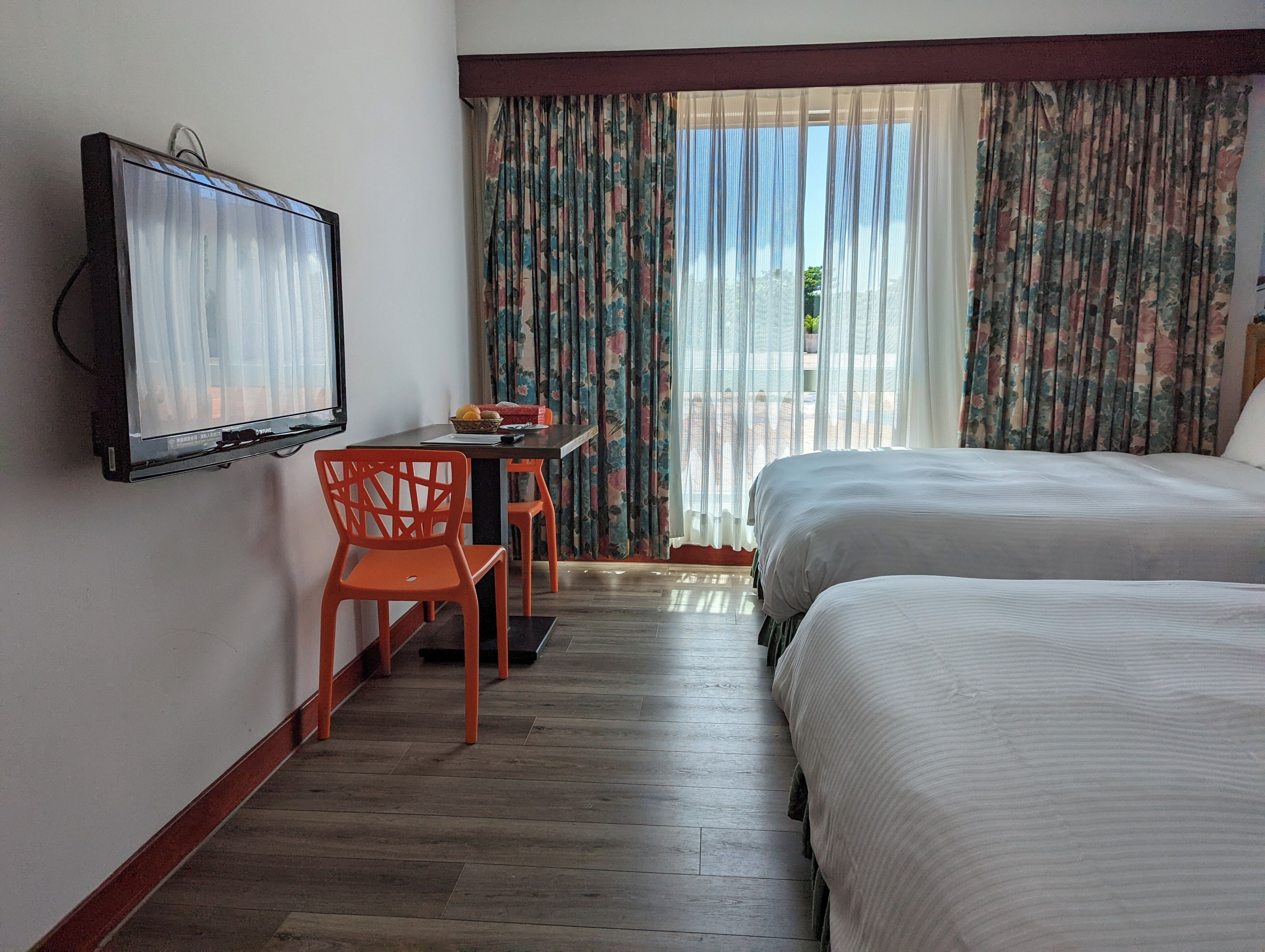 A hotel room with a bed, a TV mounted on the wall, a desk with a chair, and a window with curtains.