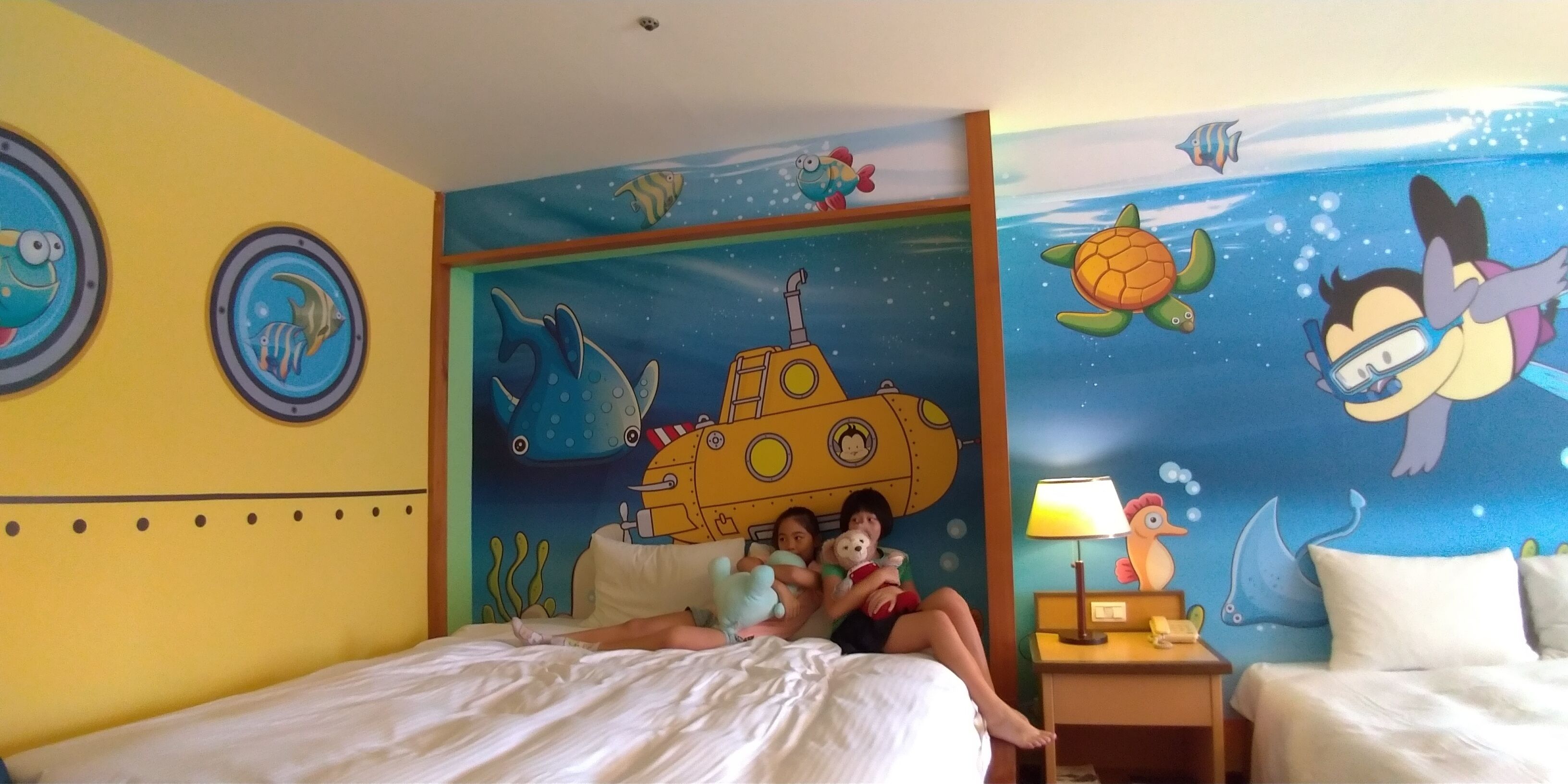 Una habitación con un mural con temática de submarino, una cama, una mesita de noche y una lámpara.