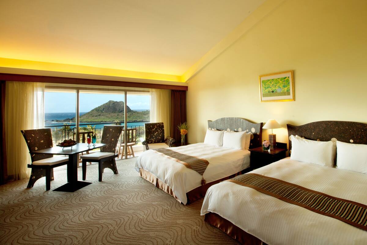 A hotel room with two beds, a dining area with a table and chairs, and a large window offering a view of the ocean and a mountain.