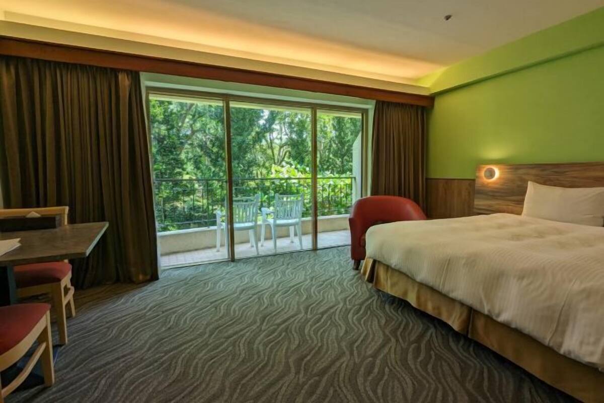 A hotel room with a bed, a desk, a chair, a balcony with a view of trees, and a patterned carpet.