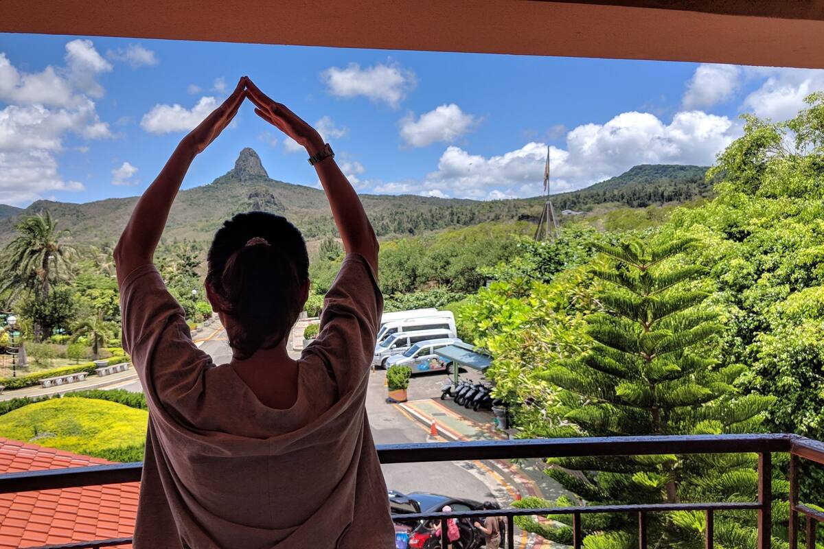 A person on a balcony with a view of a mountainous landscape, a road, and parked vehicles.