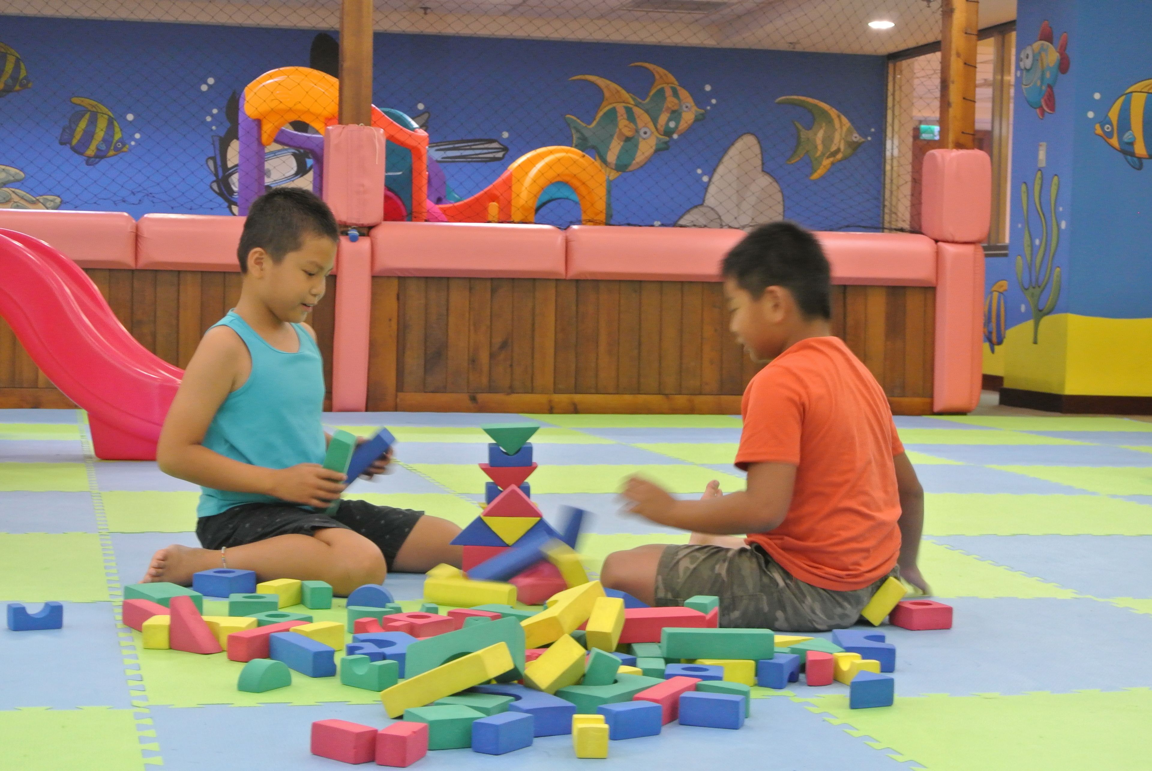 Two children playing with colorful building blocks on a mat in a room with a wooden floor and a wall mural of fish.