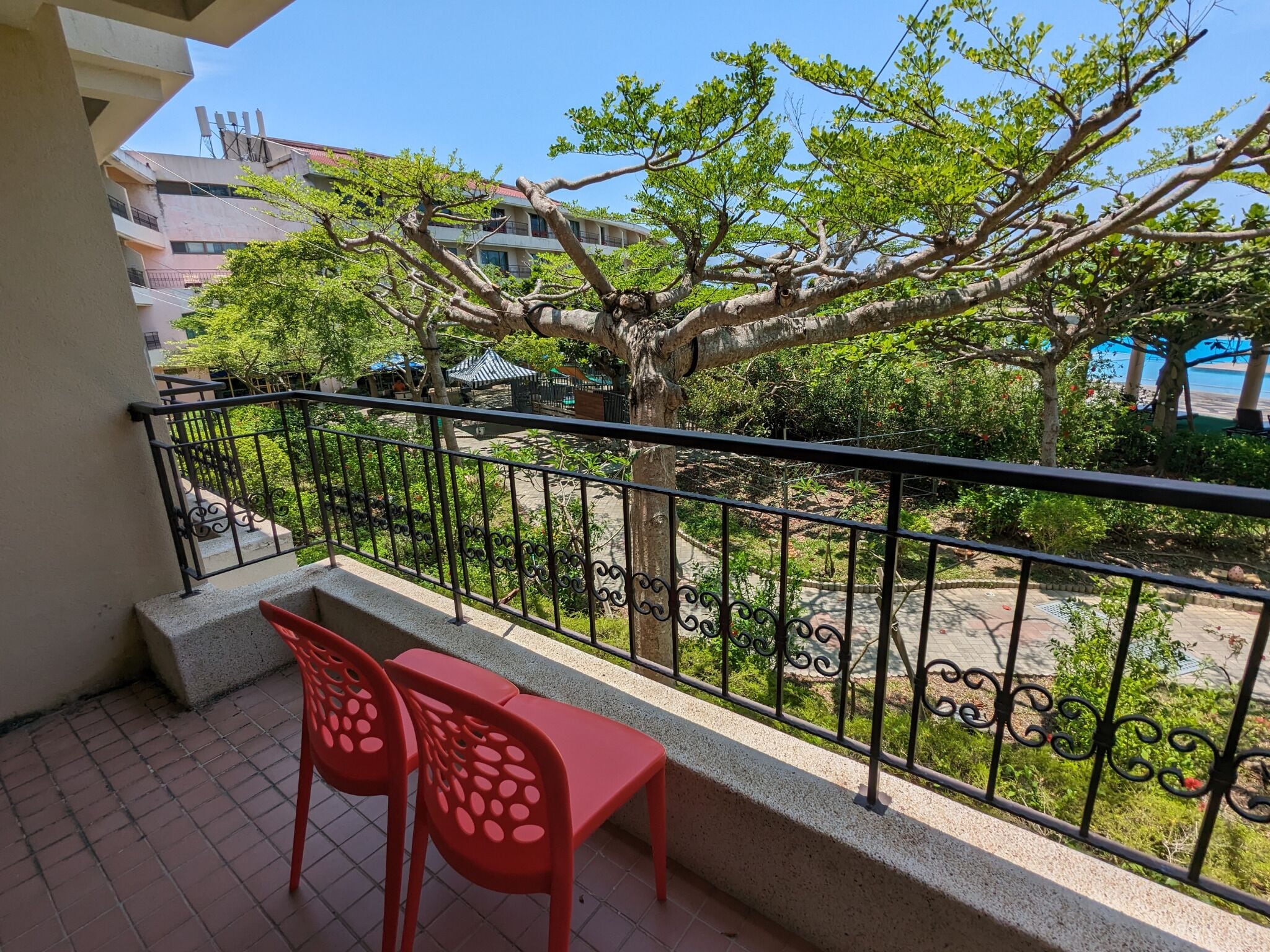 A balcony with a metal railing, two red plastic chairs, and a tree in the background.