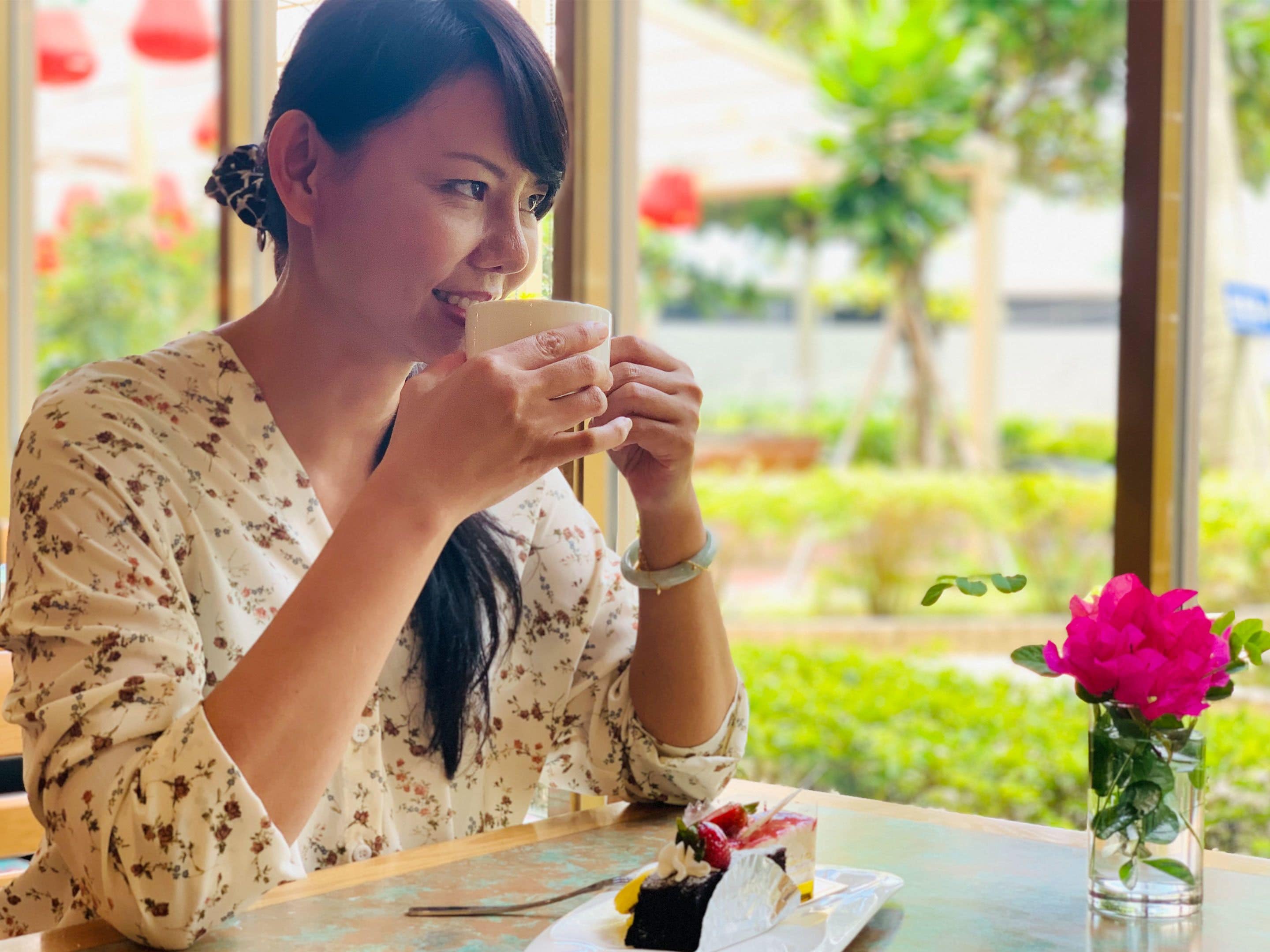 A person seated at a table with a dessert and a cup, with a window view of greenery and a pink flower in a vase.