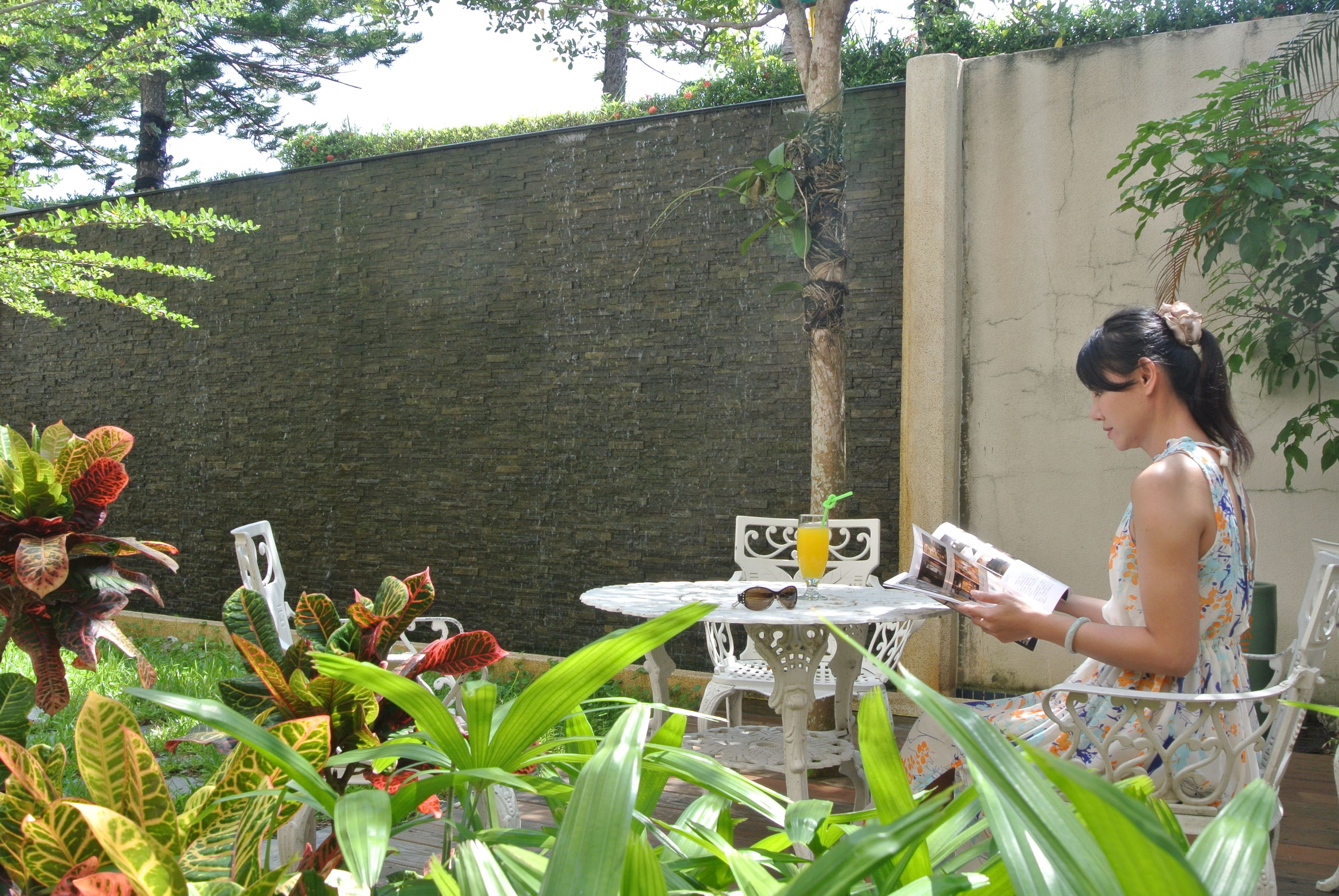 A woman seated at a white metal table with a drink, surrounded by lush green plants.