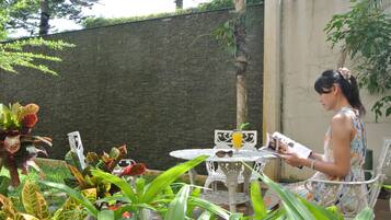 A woman seated at a white metal table with a drink, surrounded by lush green plants.
