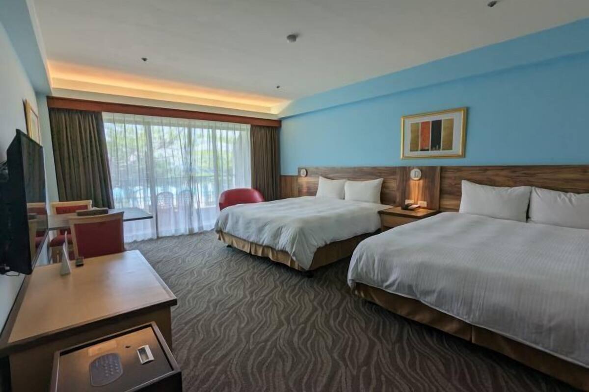 A hotel room with two beds, a desk, a TV, and a window with curtains.