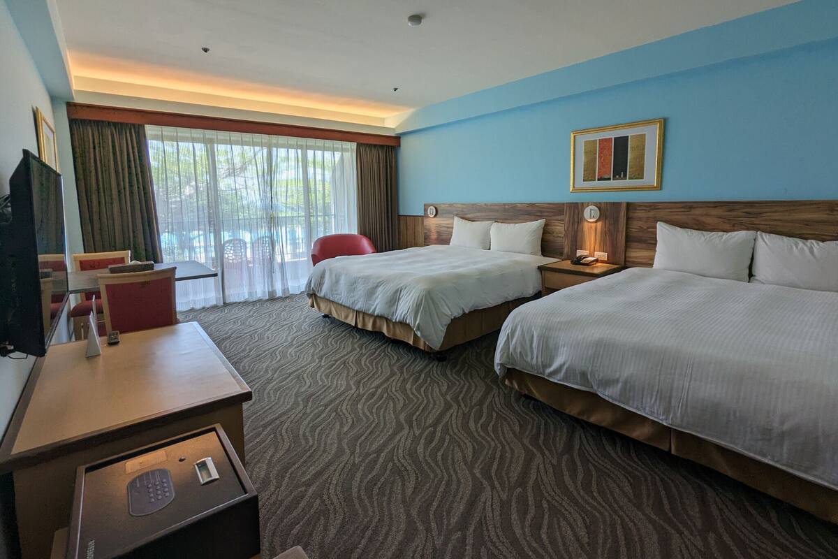 A hotel room with two beds, a desk with a telephone, a TV, and a view of the outdoors through large windows.