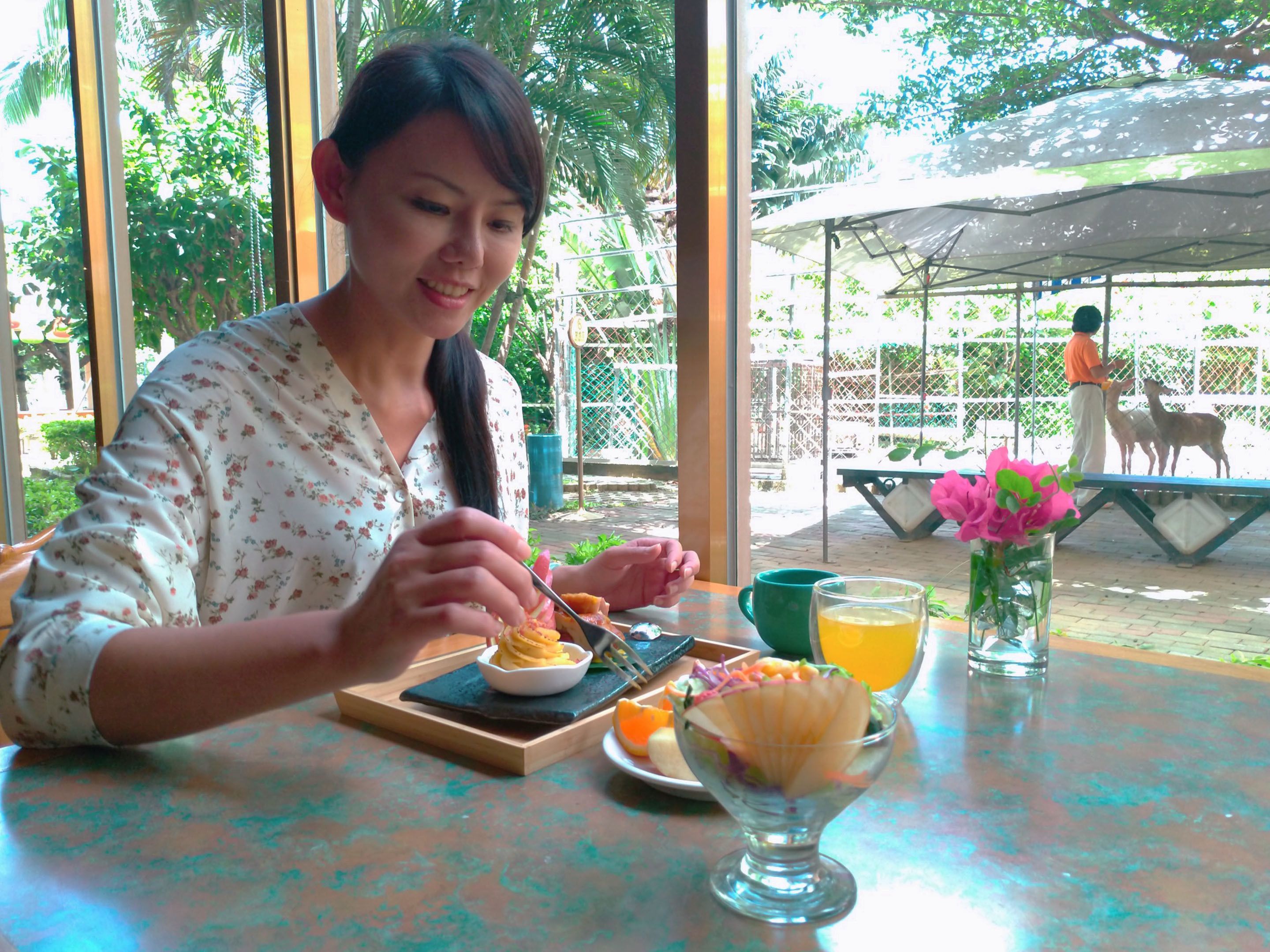 A person dining at an outdoor table with a meal, a drink, and a flower vase.