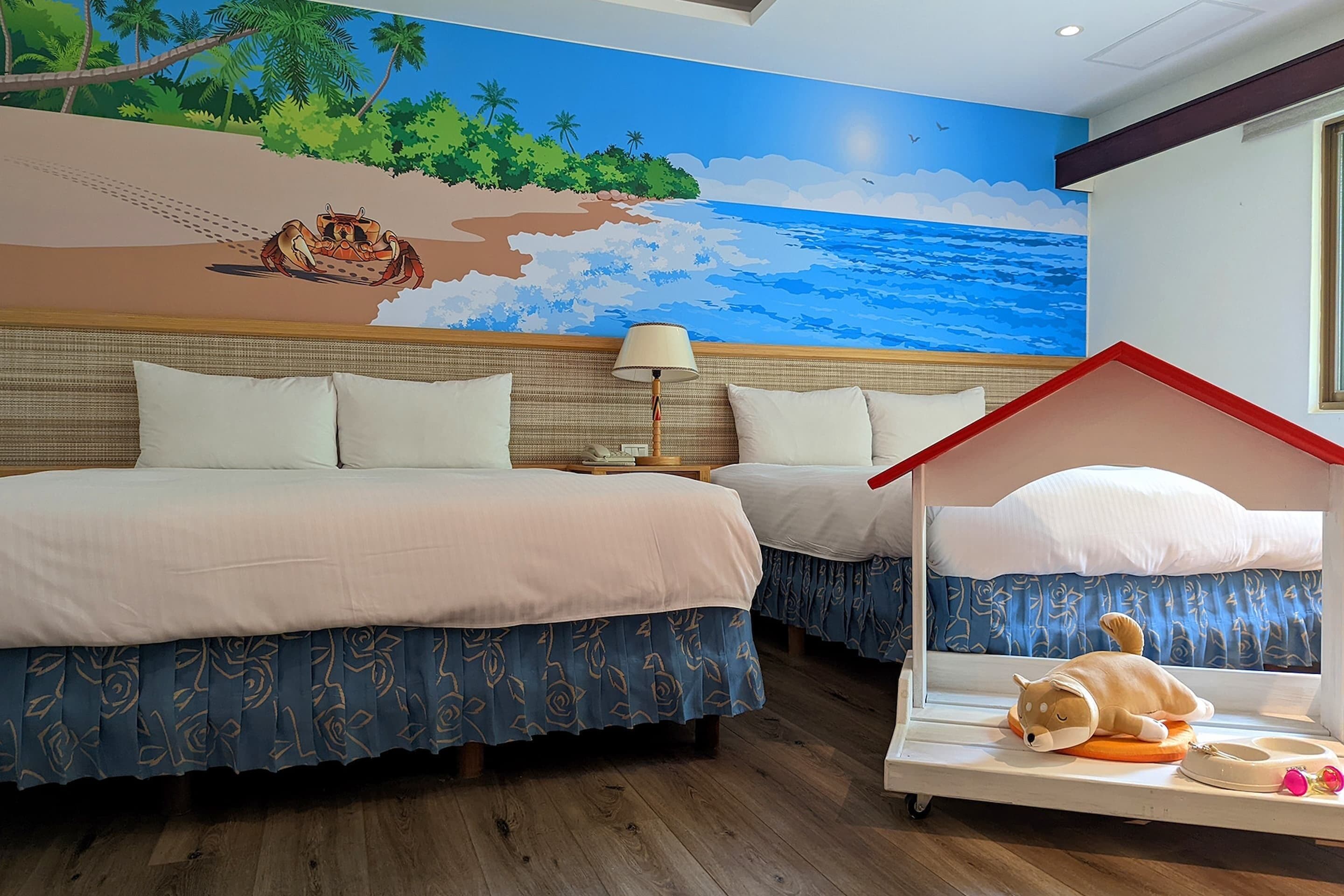 Hotelkamer met twee bedden, een muurschildering van een strand en een lamp, plus een klein hondenbed met speeltje.