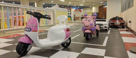 Un espace de stationnement avec des scooters et des voitures, un sol à carreaux et, au fond, une structure de jeu colorée.