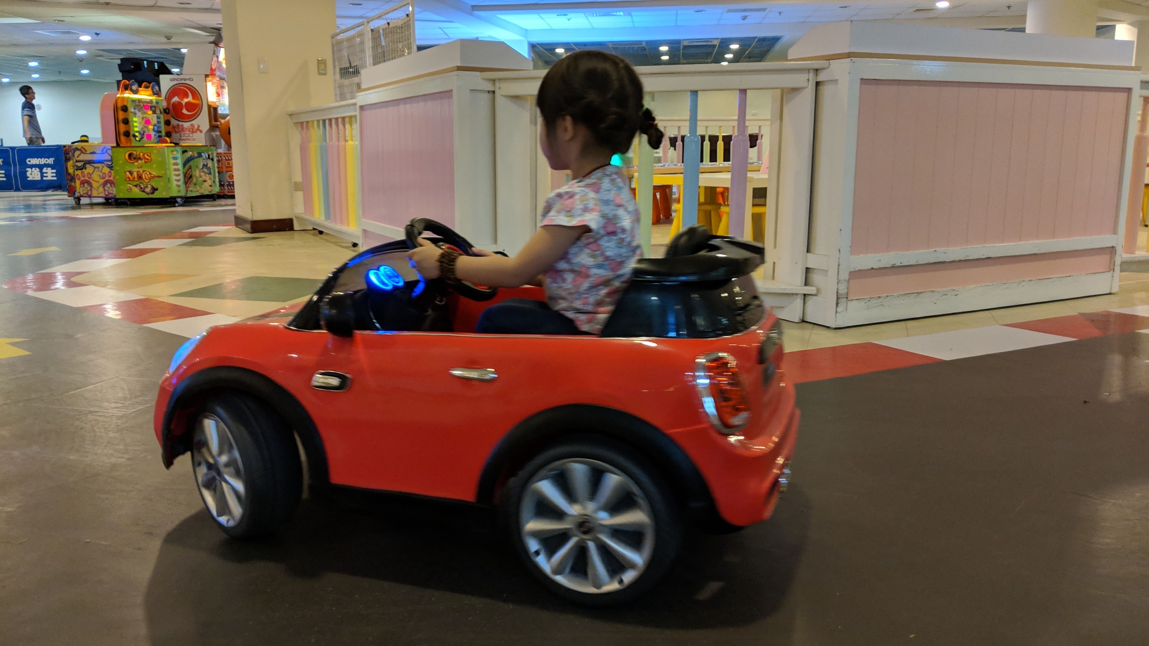 Un niño conduce un coche de juguete rojo en un área de juegos interior con estructuras de juego coloridas y un suelo a cuadros.