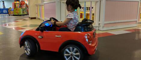 A child driving a red toy car in an indoor play area with colorful play structures and a checkered floor.