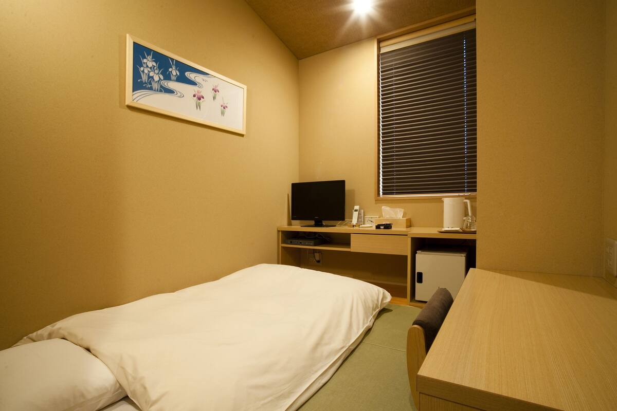 Camera d'albergo con due letti singoli, una scrivania con computer, un piccolo frigorifero e una finestra con persiane.