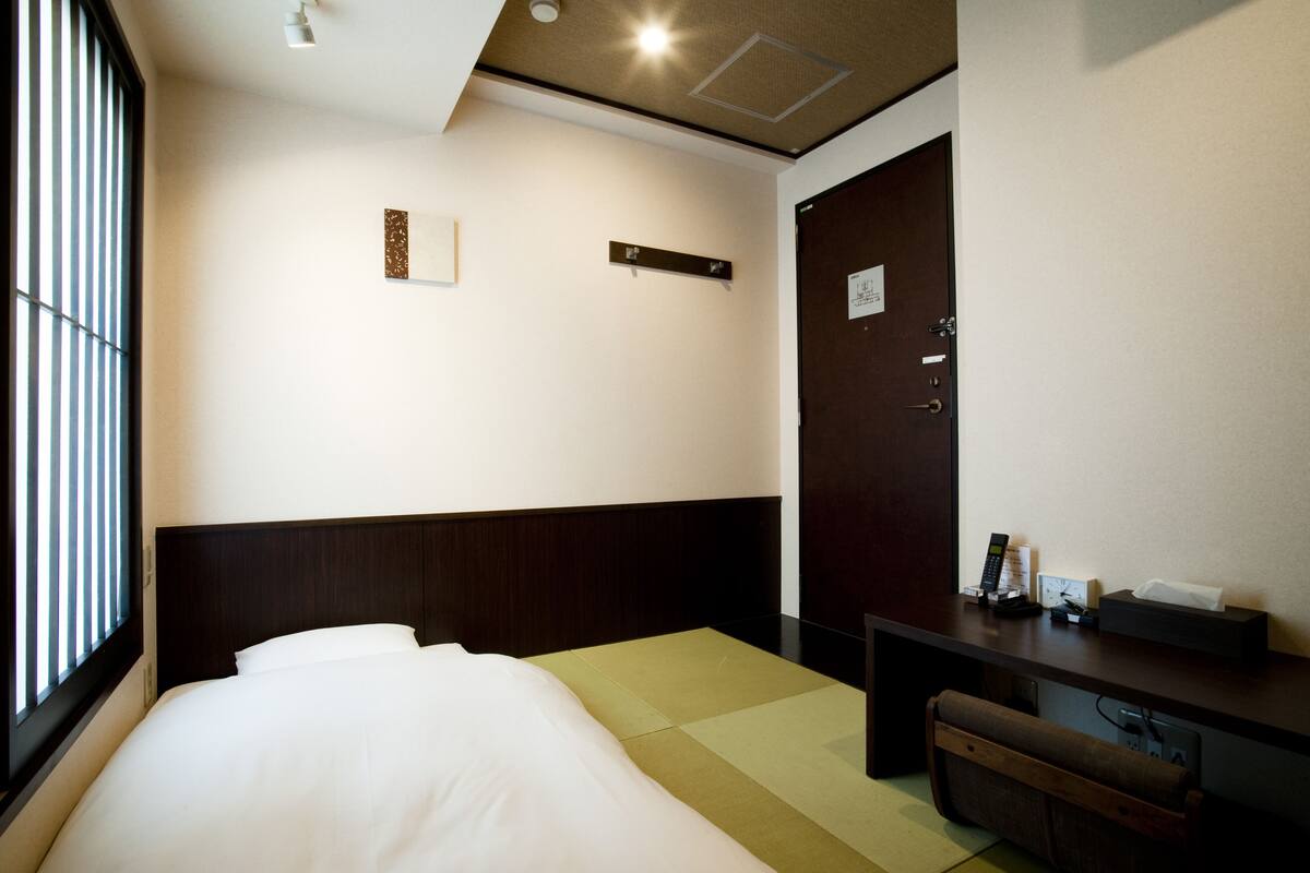 Una camera d'albergo con un letto, una scrivania, una sedia, un comodino e una porta.