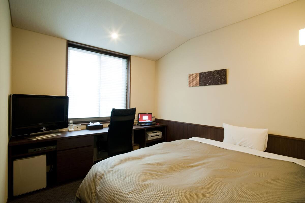 Una camera d'albergo con un letto, una scrivania con un computer, una televisione e una finestra.