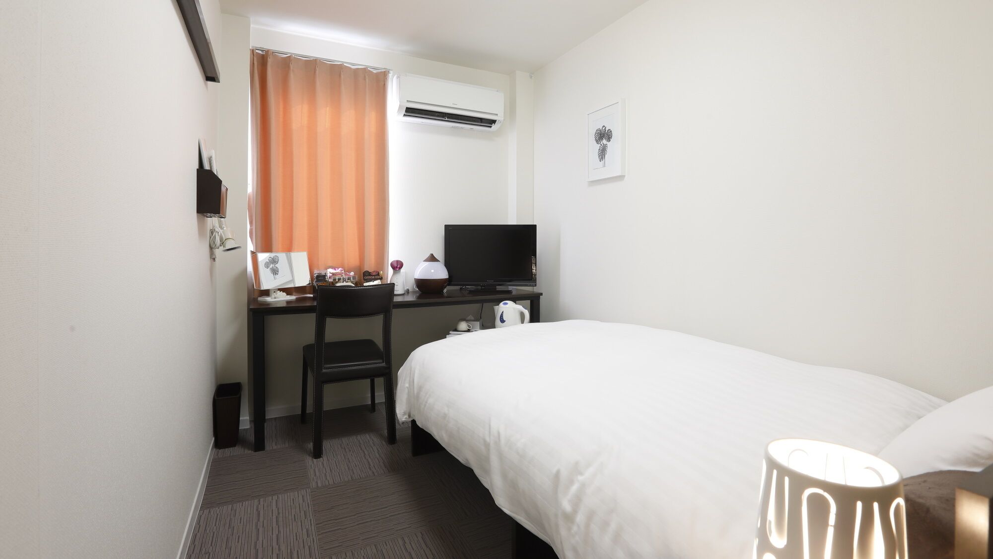 A hotel room with a bed, a desk with a computer, a chair, a TV, and an air conditioning unit.