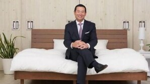 A man sitting on a bed in a hotel room with a wooden headboard, a potted plant, and a bedside table with a lamp. - Value The Hotel Sendai Natori (Natori)