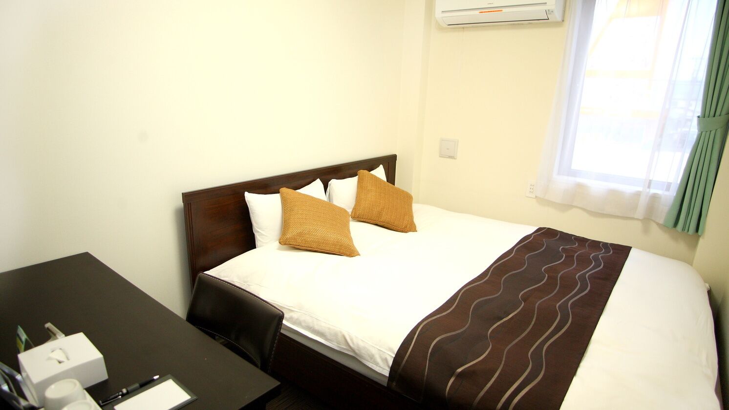 A hotel room with a bed, a desk with a chair, a window with curtains, and an air conditioning unit.