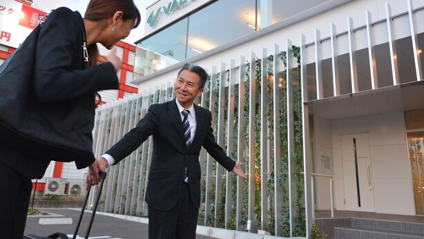 A man in a suit with a briefcase standing in front of a building with a sign that reads "VALUE THE". - Value The Hotel Sendai Natori (Natori)