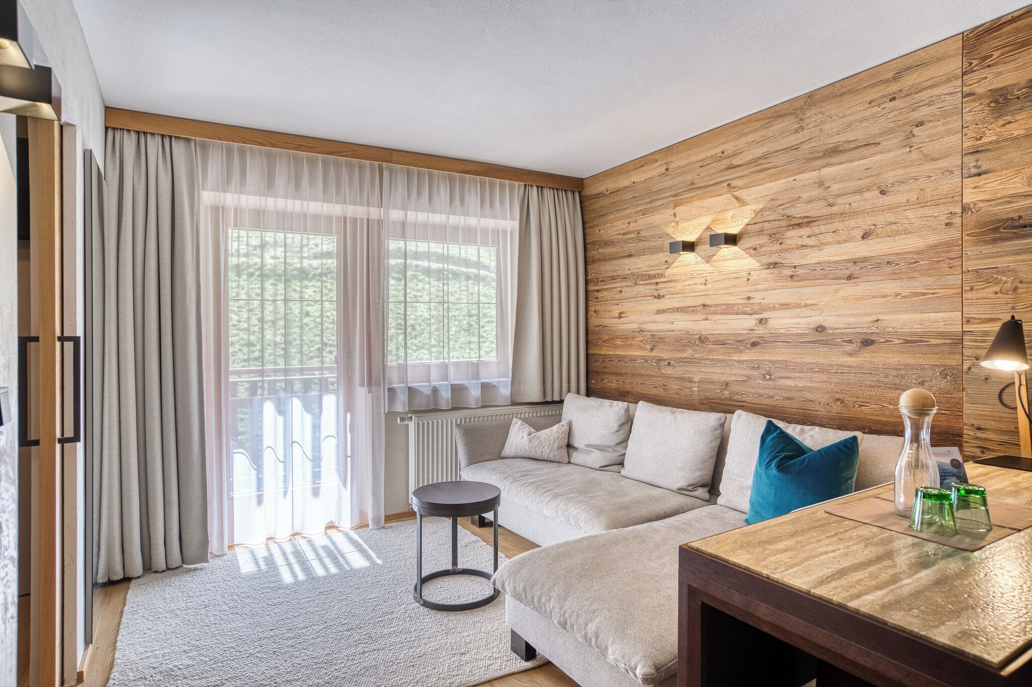 A modern living room with a wooden wall panel, a sofa, a round table, and a large window with curtains.