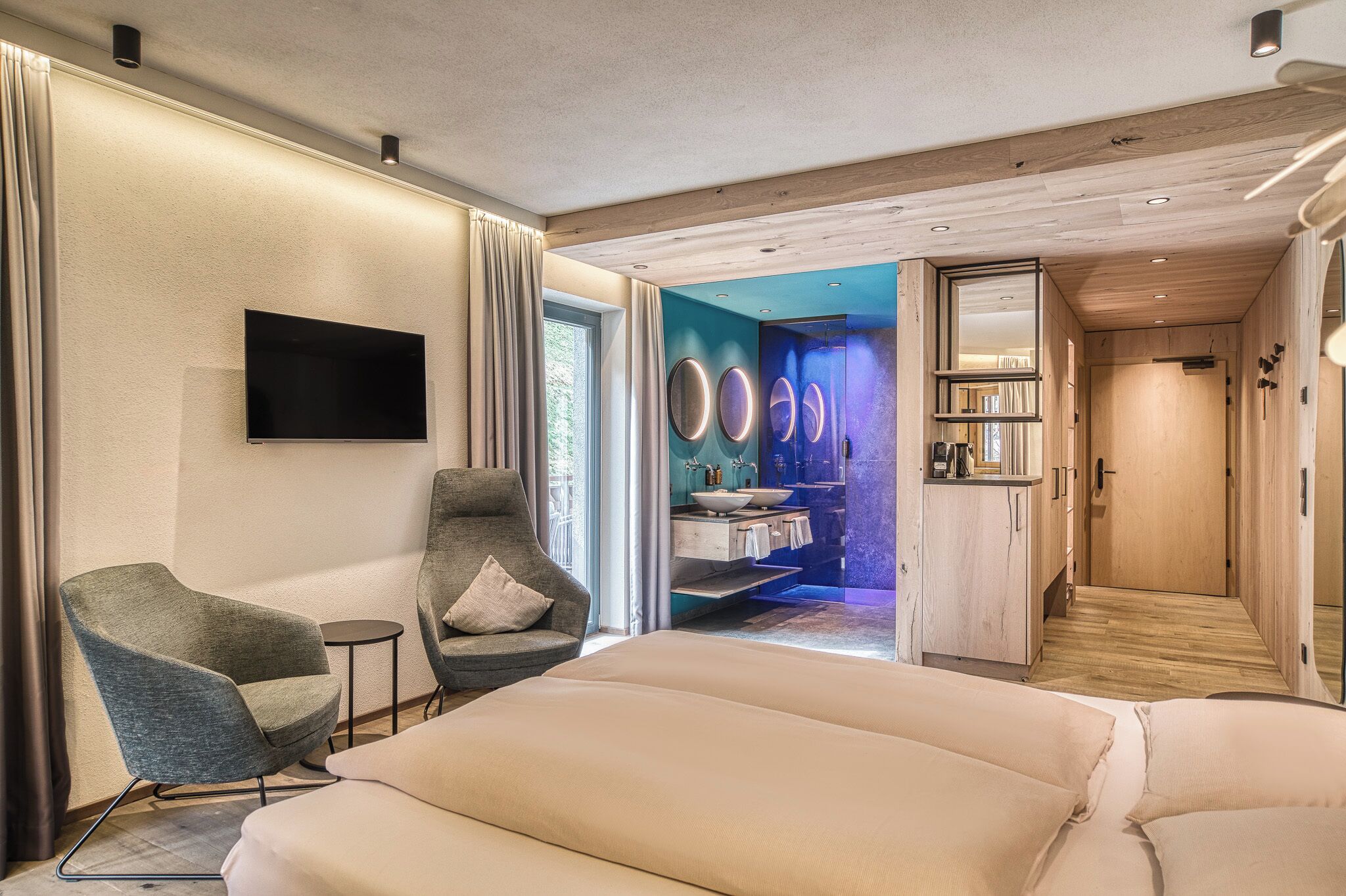 A modern hotel room with a large bed, two armchairs, a TV, and a bathroom with a vanity and mirror.