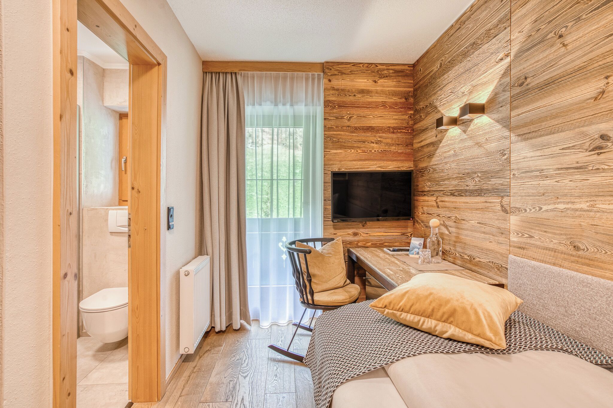 A compact room with a wooden wall, a television, a rocking chair, a sofa, and a window with curtains.