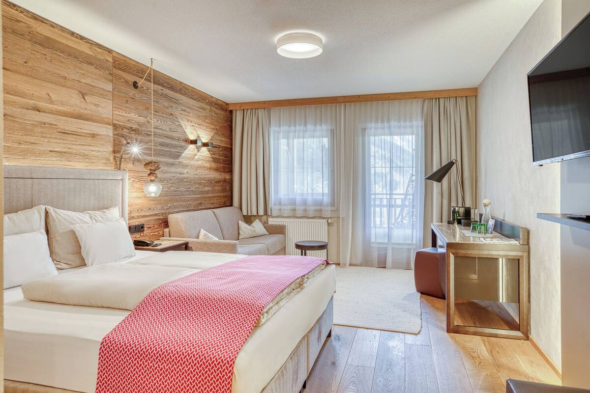 Ein modernes Hotelzimmer mit einer Wand aus Holzvertäfelung, einem großen Bett, einem Schreibtisch, einem Fernseher und einem Fenster mit Vorhängen.