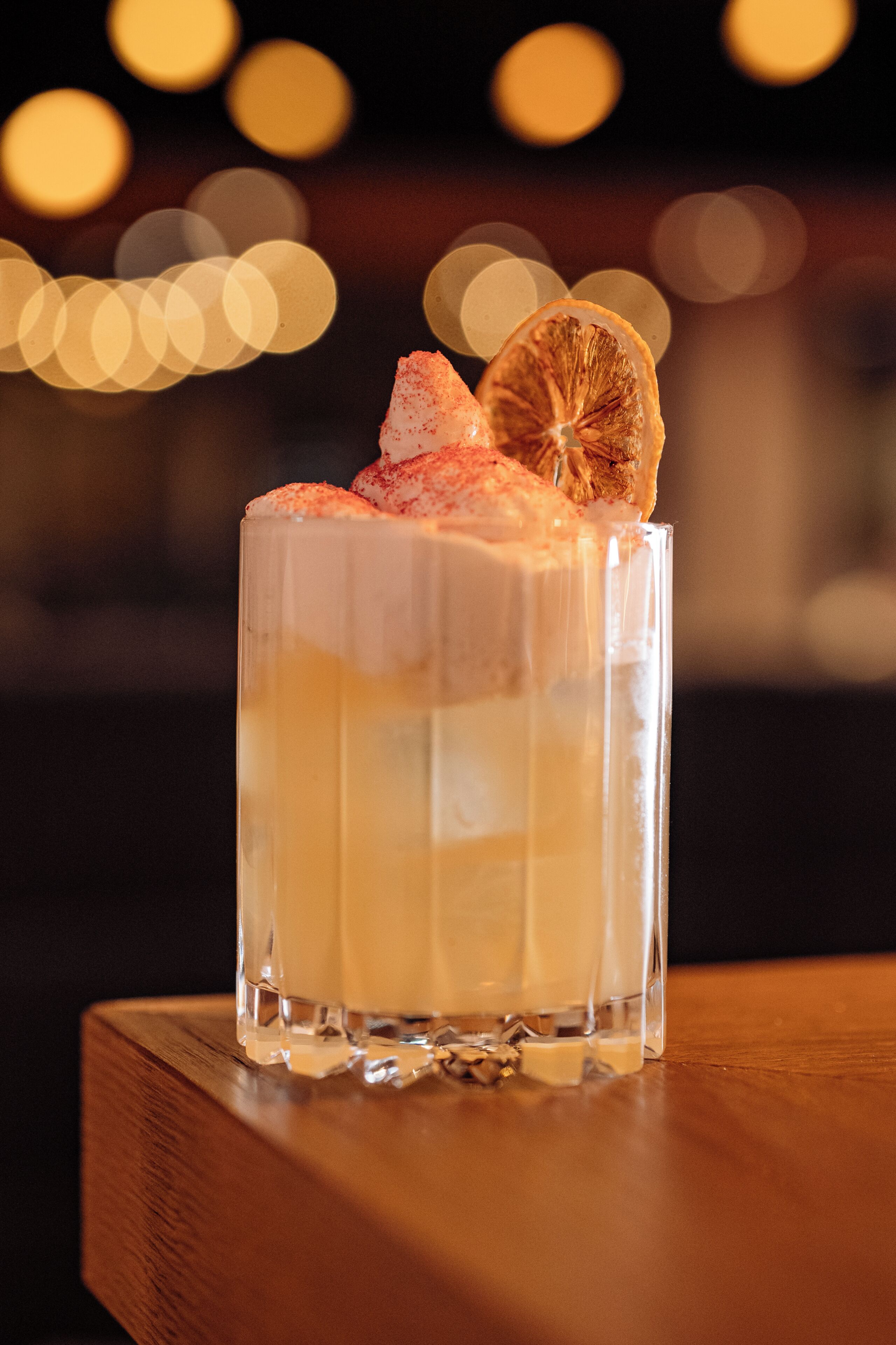 Un vaso transparente con una bebida en capas, adornado con una rodaja de naranja y un poco de condimento, sobre una superficie de madera.