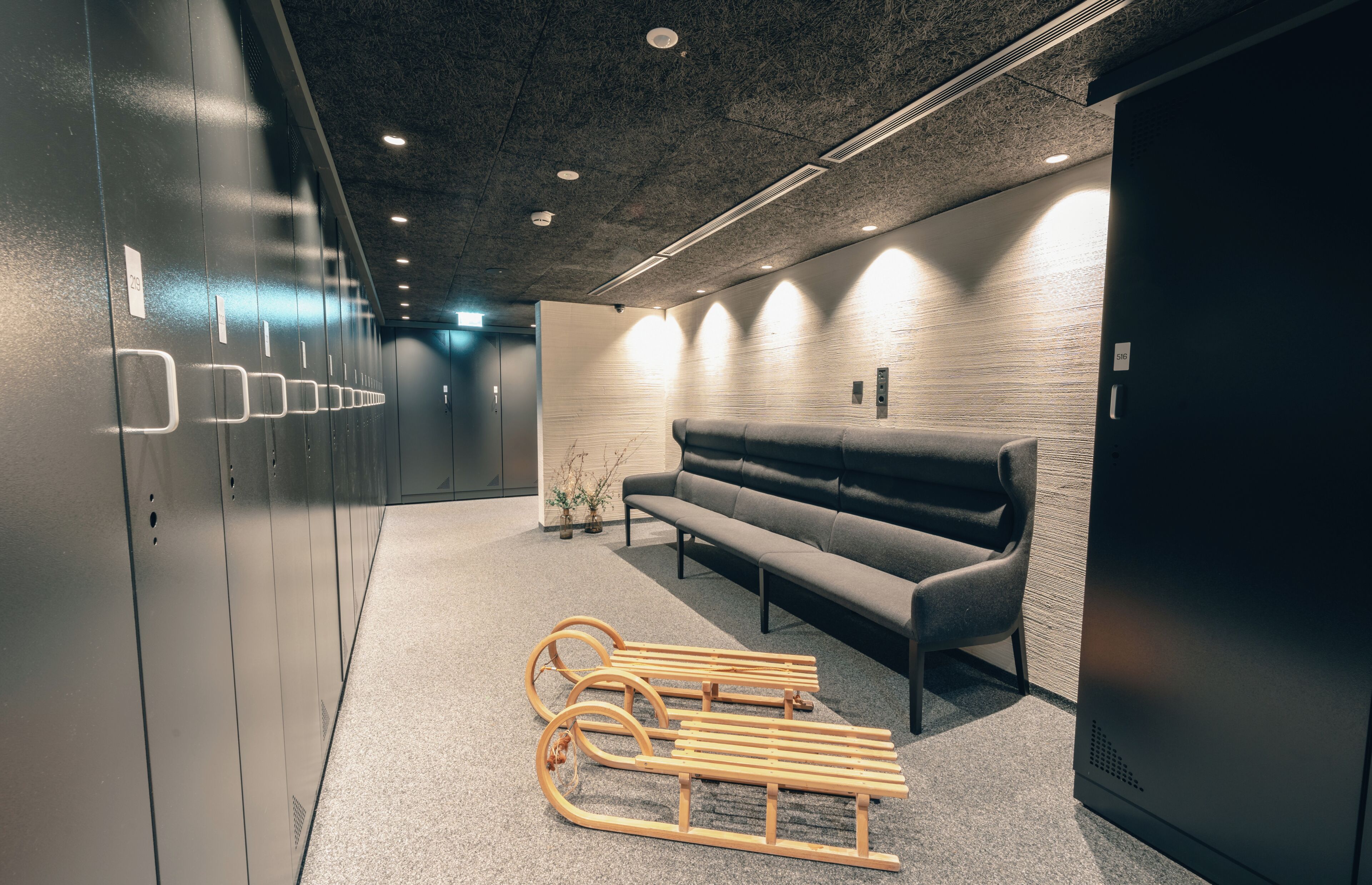 A modern waiting area with a sofa, wooden footrest, and a wall with lockers.