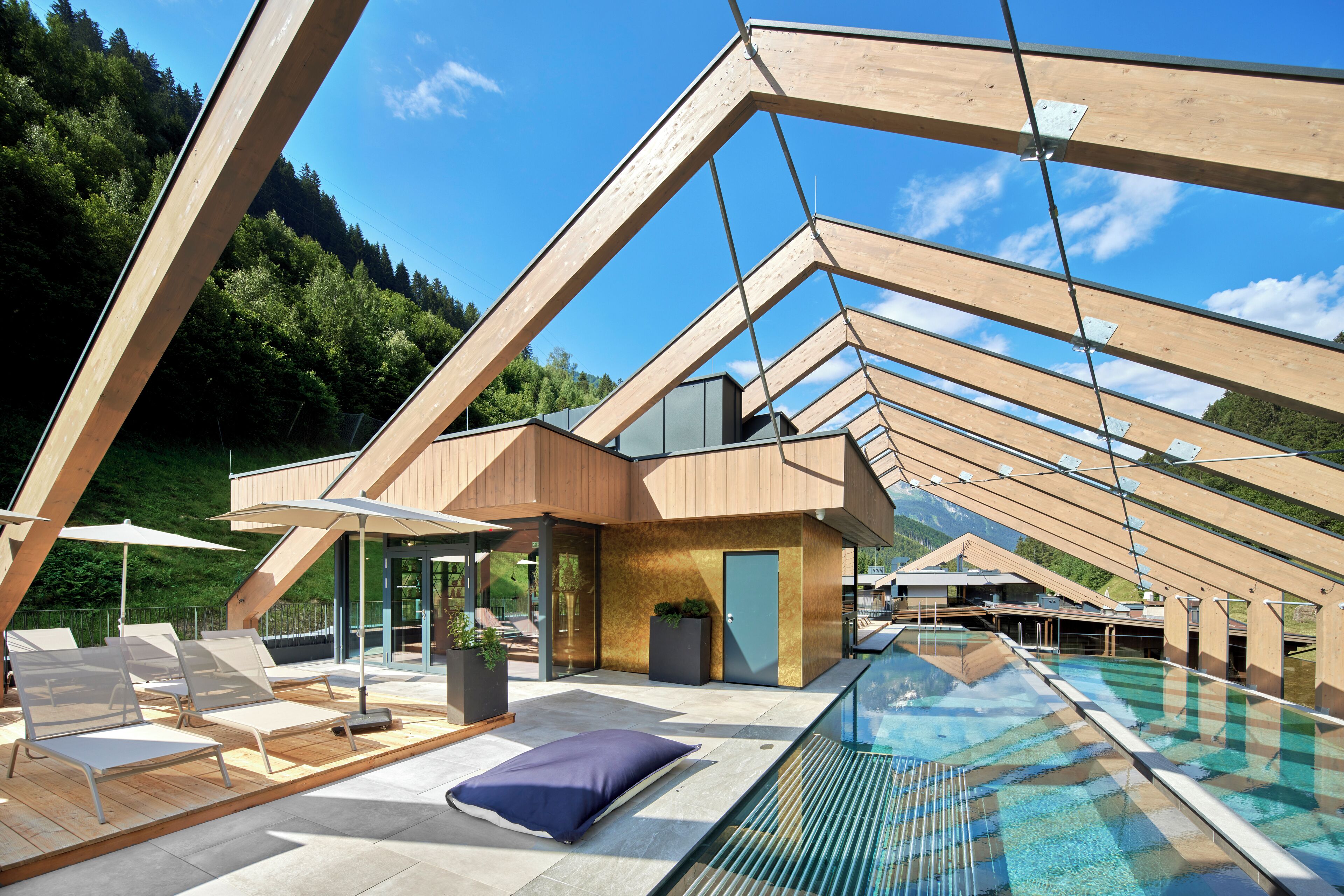 Un área de piscina exterior moderna con vigas de madera, sillones de descanso y una piscina cubierta de cristal.