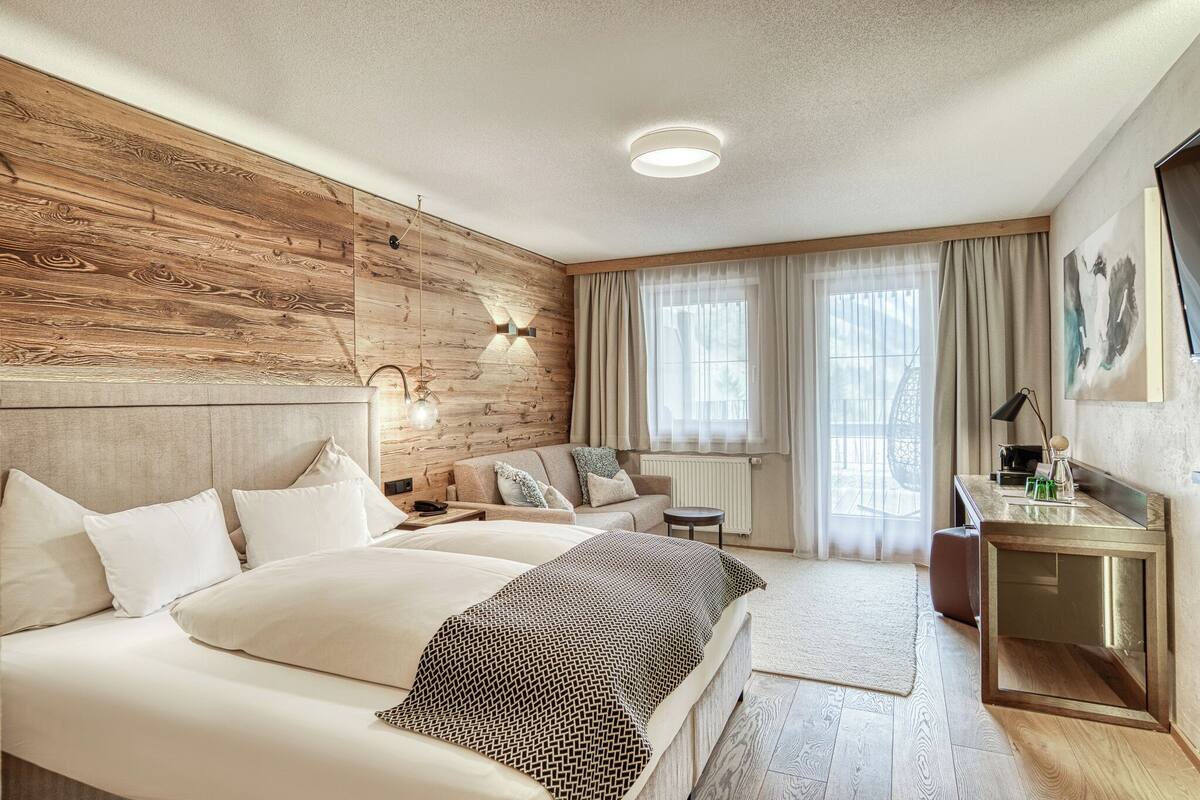 Ein Hotelzimmer mit einer Wandpaneele aus Holz, einem Bett mit weißer Bettwäsche, einem Schreibtisch mit Spiegel und einem großen Fenster mit durchsichtigen Vorhängen.