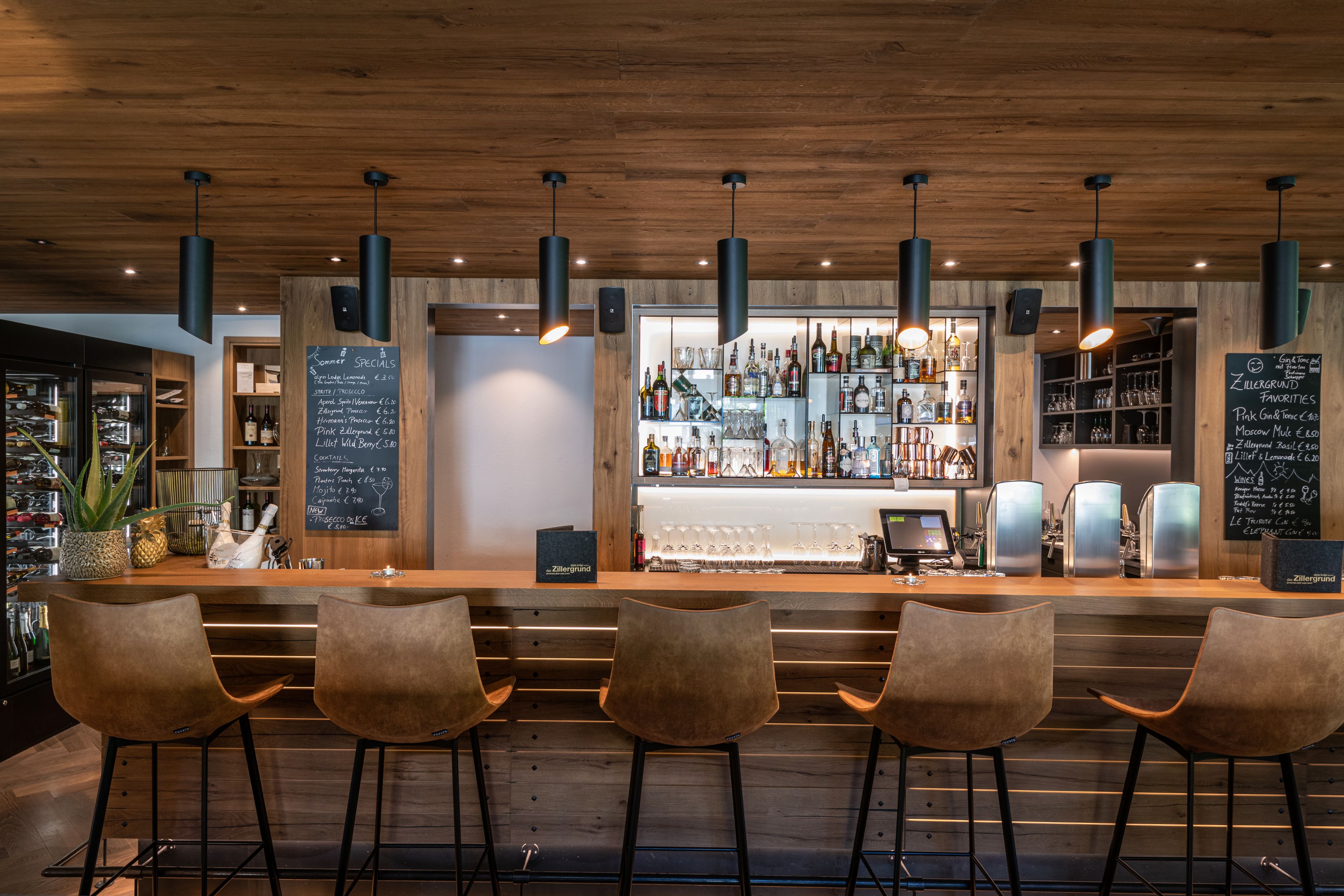 A bar area with wooden ceiling, hanging pendant lights, and bar stools. There are shelves with bottles and glasses behind the bar.