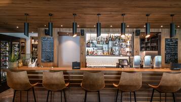 A bar area with wooden ceiling, hanging pendant lights, and bar stools. There are shelves with bottles and glasses behind the bar.