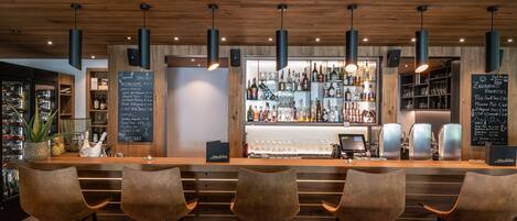 A bar area with wooden ceiling, hanging pendant lights, and bar stools. There are shelves with bottles and glasses behind the bar.