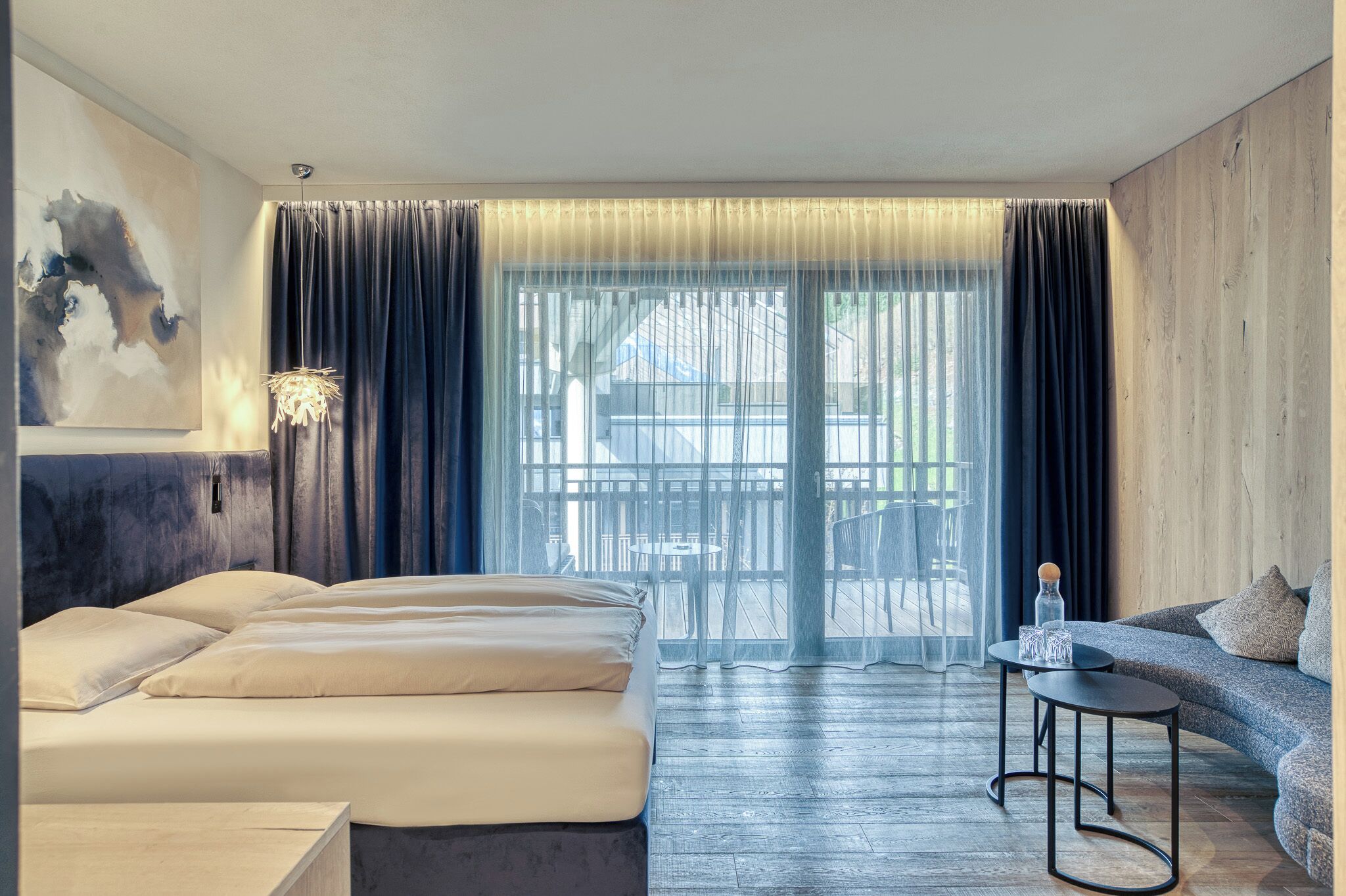 Ein Hotelzimmer mit einem Bett, einem Sofa, einem kleinen Tisch und Blick durch ein großes Fenster mit Vorhängen.