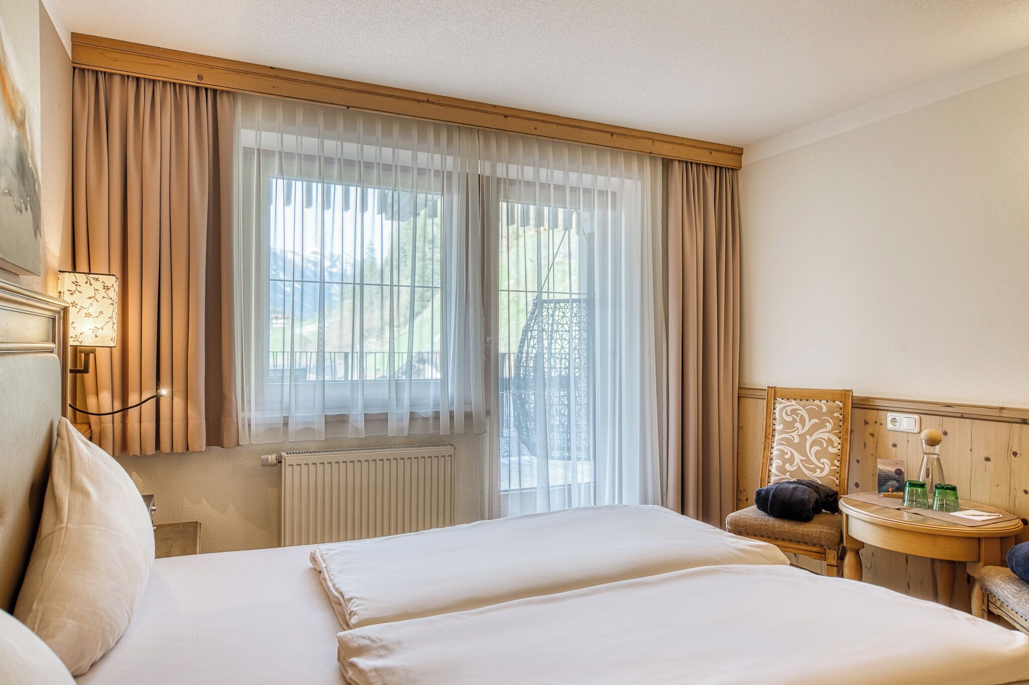 Une chambre d’hôtel avec un grand lit, une petite table avec une lampe, une chaise et une fenêtre avec des rideaux légers.