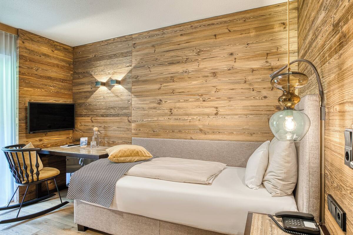 Ein modernes Zimmer mit Holzpaneelen, einem Flachbildfernseher, einem Bett mit Kissen, einem Schreibtisch mit Stuhl und einem Telefon.