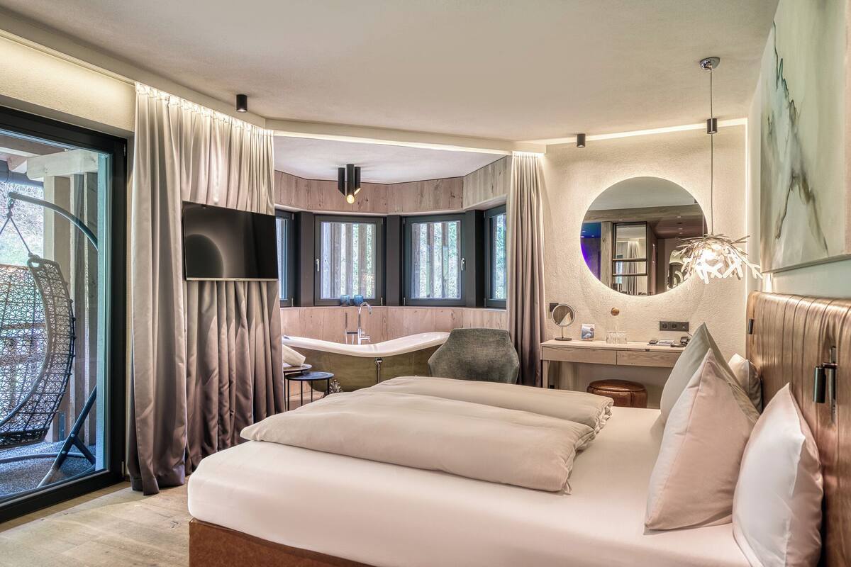 Ein modernes Hotelzimmer mit einem großen Bett, einem Schreibtisch, einem Sessel, einer Badewanne und einem großen Fenster.