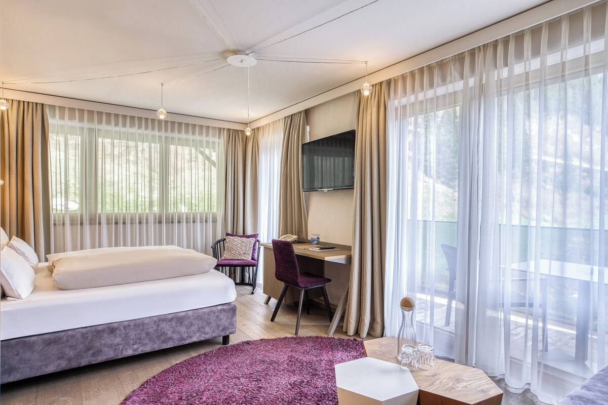 Ein Hotelzimmer mit Bett, Schreibtisch, Stuhl, Fernseher und großen Fenstern mit Vorhängen.