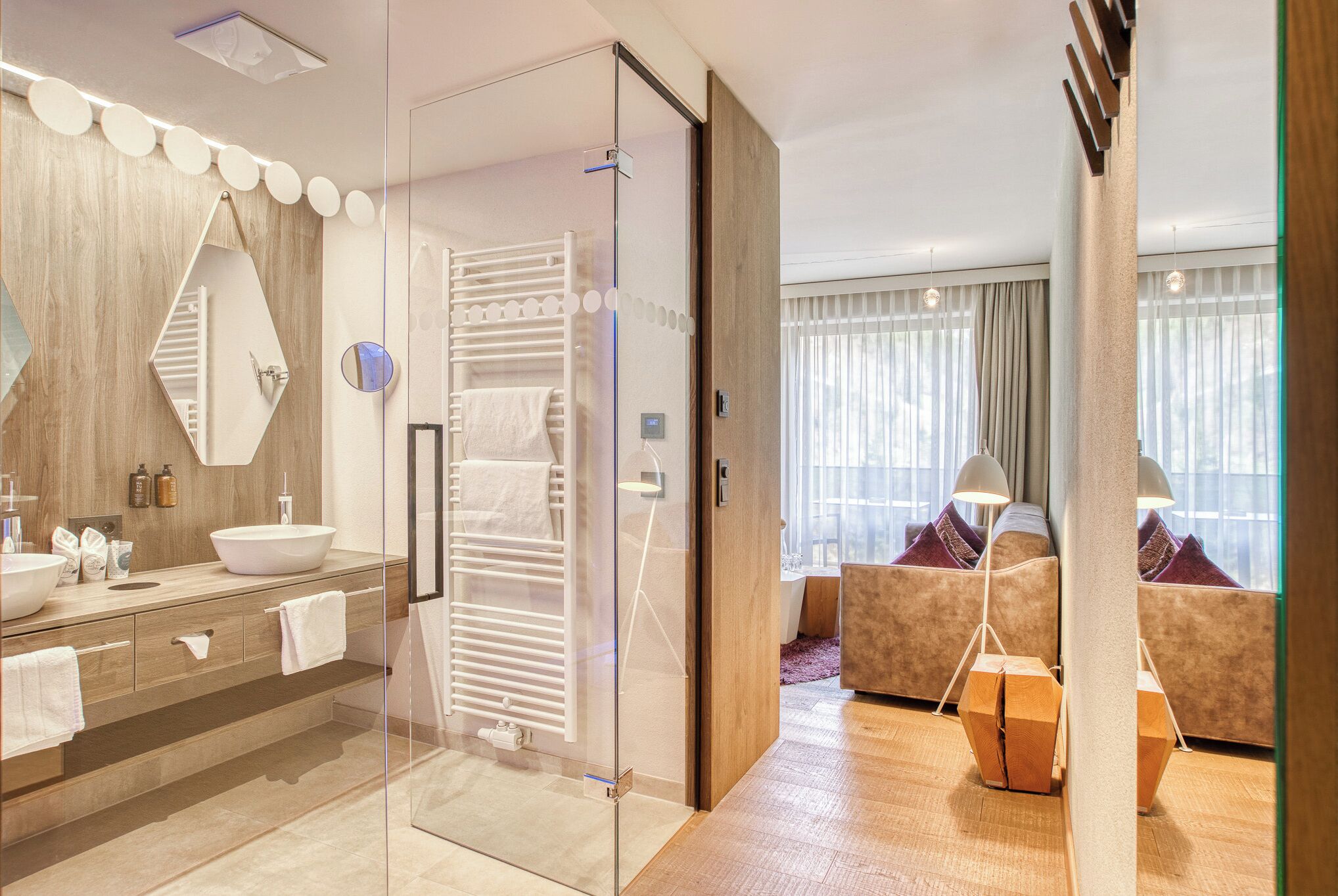 A modern hotel room with a glass-enclosed shower, a large mirror, a wooden vanity with a sink, and a seating area with a sofa and cushions.