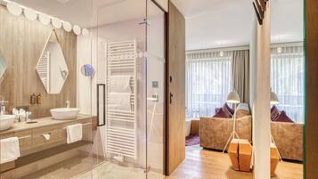A modern hotel room with a glass-enclosed shower, a large mirror, a wooden vanity with a sink, and a seating area with a sofa and cushions.