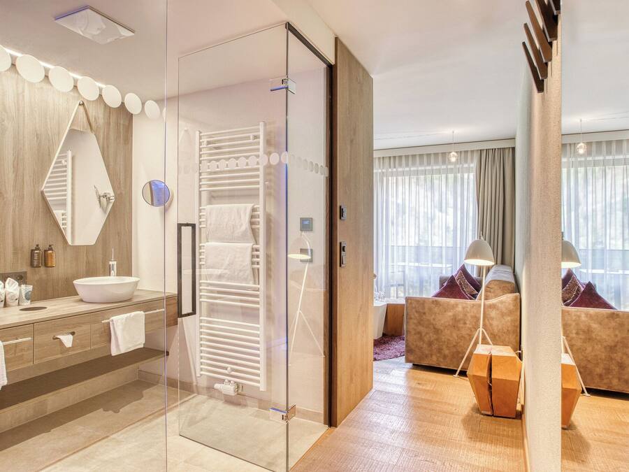 A modern hotel room with a glass-enclosed shower, a large mirror, a wooden vanity with a sink, and a seating area with a sofa and cushions.
