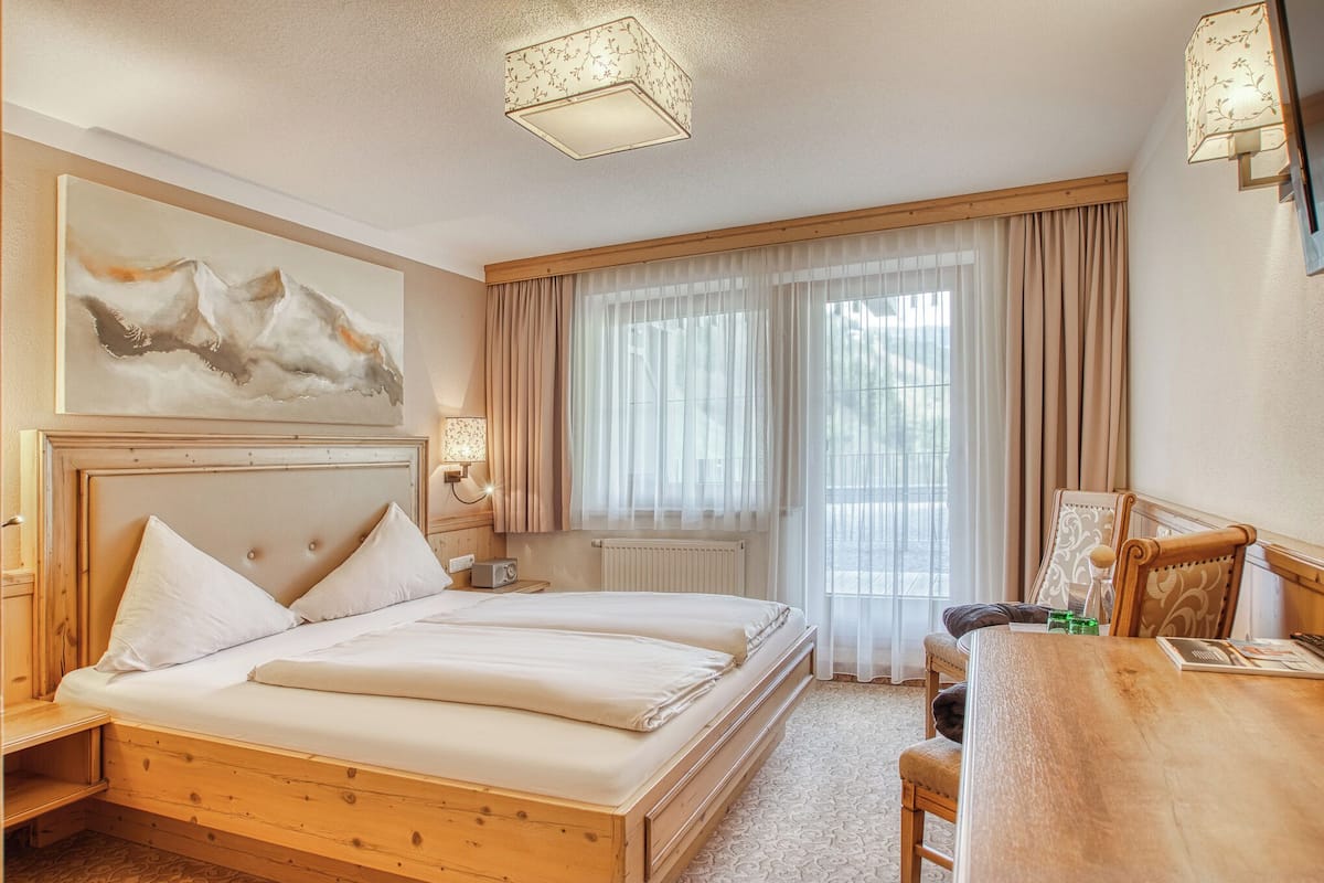 Ein Hotelzimmer mit einem hölzernen Bett, einem Schreibtisch, einem Sessel, einem großen Fenster mit leichtem Vorhang und einem Bild an der Wand.