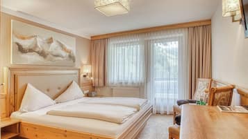 A hotel room with a wooden bed, a desk, a chair, a large window with sheer curtains, and a painting on the wall.