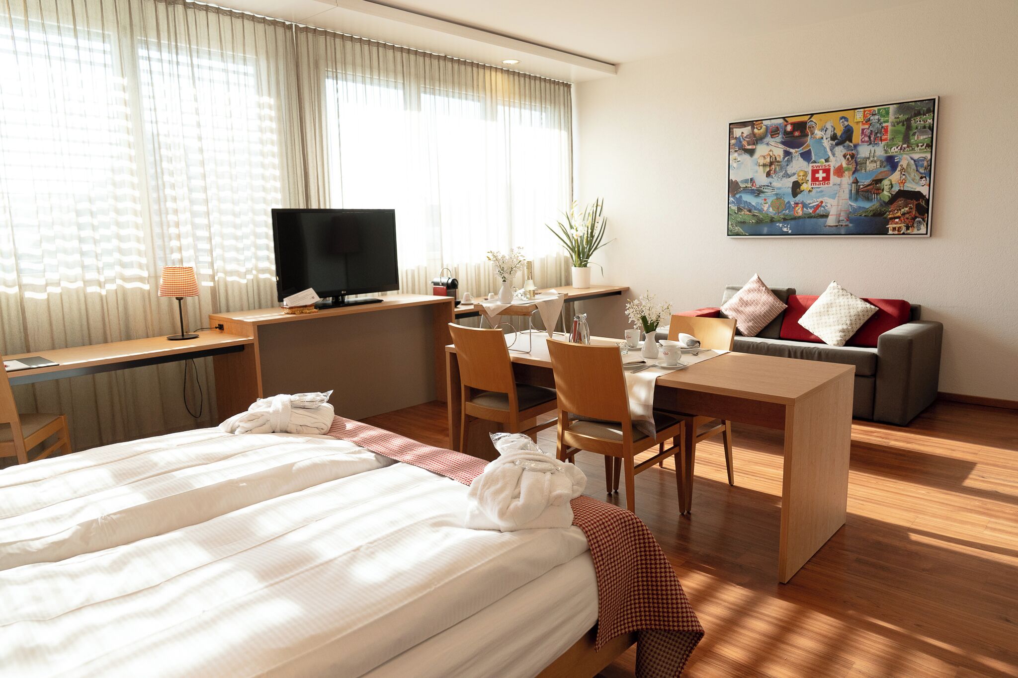 Una habitación de hotel con una cama, un escritorio con televisor, una mesa de comedor con sillas, un sofá y un cuadro en la pared.