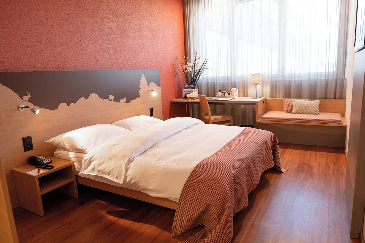 A hotel room with a bed, bedside table, lamp, and a small desk with a chair.