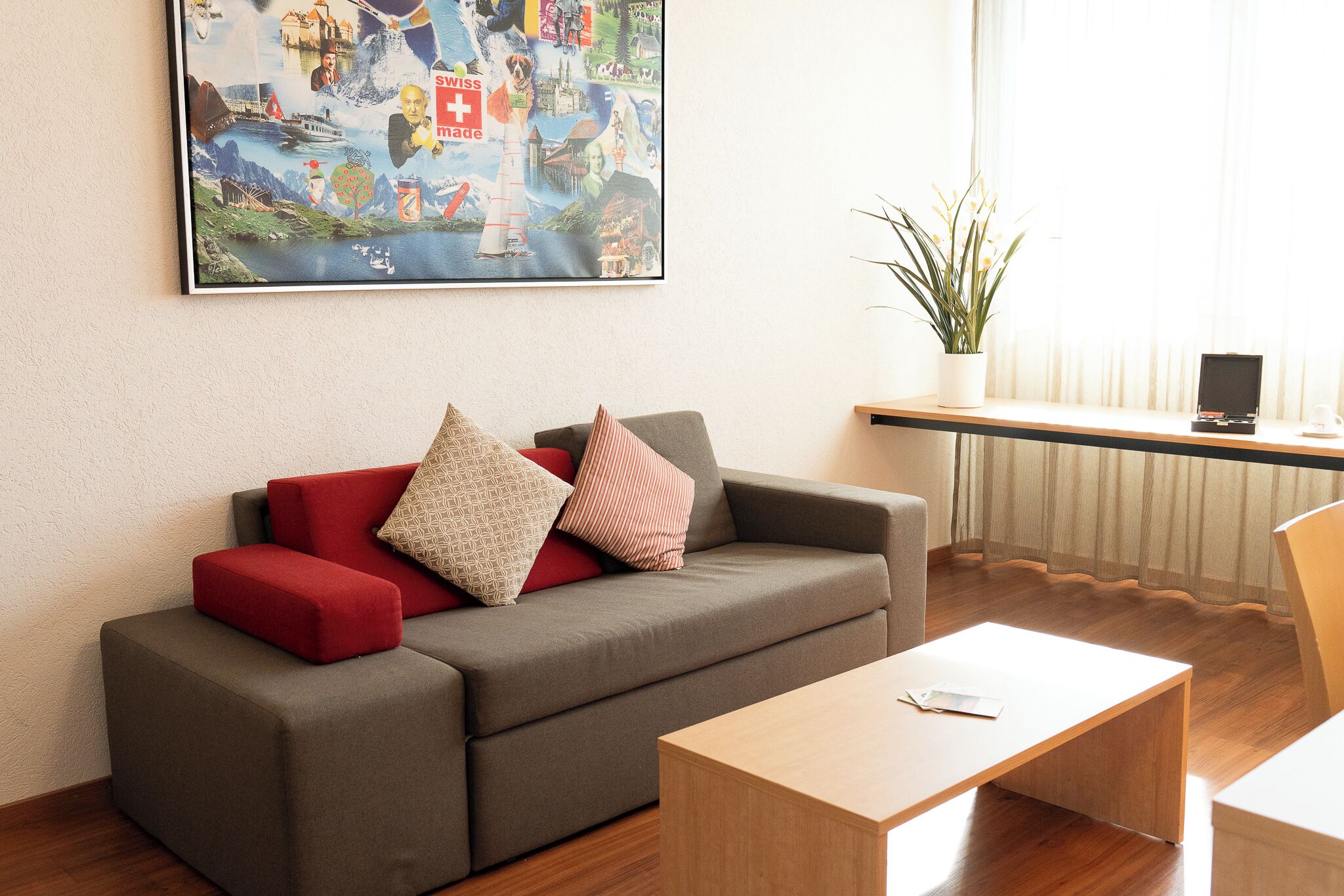 A modern living room with a grey sofa, red and patterned cushions, a wooden coffee table, and a large poster on the wall.